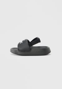 Μαύρο σανδάλι slides από καουτσούκ με ελαστικό λουράκι πίσω. Διαθέτει υφασμένο πέλμα και λευκό λογότυπο Lacoste στο πάνω μέρος.