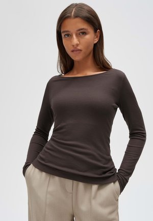 Junge Frau mit langen braunen Haaren, die ein dunkelbraunes Langarmoberteil und beige Hose trägt, mit den Händen in den Taschen, neutraler Hintergrund.