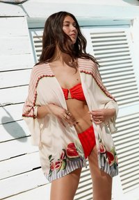 Lagani, proziran kimono s cvjetnim uzorcima, crvenim detaljima i prugastim rukavima. Nosite ga preko crvenog bikinija, prikazujući opušteni stil plaže.