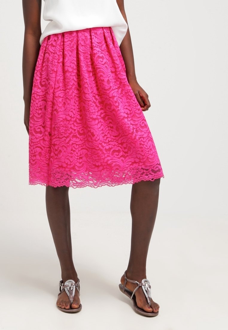 Kobieta w różowej koronkowej spódnicy do kolan, białej bluzce i srebrnych płaskich sandałach stoi na jednolitym jasnym tle.