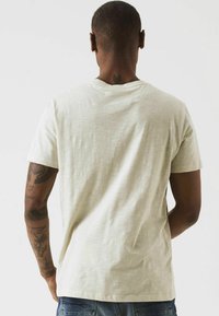 Camiseta de manga corta color beige claro, hecha de tela suave, con cuello redondo y una textura sutil. Tatuajes visibles en los brazos.