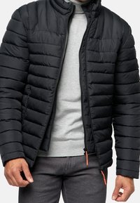 Schwarze Pufferjacke mit horizontalem Steppdesign, hohem Kragen und Reißverschluss vorne. Mit orangefarbenen Reißverschluss-Ziehern und grauem Innenfutter.