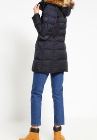 Gewatteerd jack in donker marineblauw, met een capuchon gevoerd met nepbont, doorgestikt ontwerp en zijzakken, gecombineerd met een blauwe spijkerbroek en tankleurige laarzen.