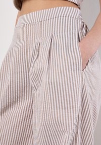 Pantalons en coton rayés marron clair et blanc avec des plis, présentant une texture lisse et des poches latérales, conçus pour le confort et une tenue décontractée.
