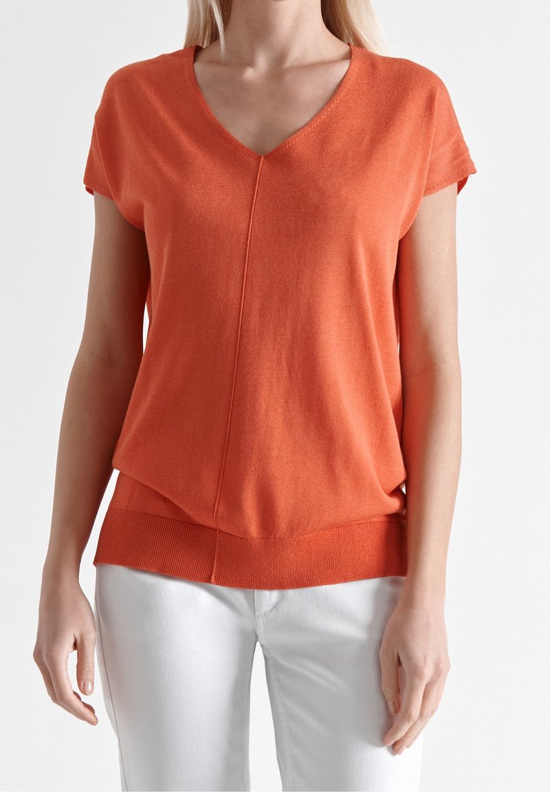 Top de punto naranja con escote en V y mangas cortas. Presenta un corte holgado, dobladillo acanalado y una superficie ligeramente texturizada. Se lleva con pantalones blancos.