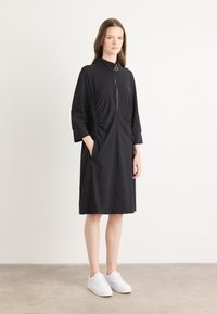 Zwarte jurk met hoge kraag, ritssluiting aan de voorkant en zijzakken. Heeft gatherdetails in de taille en driekwart mouwen.