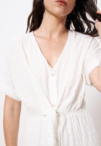 Weißes, zerknittertes Kleid aus Stoff mit V-Ausschnitt, kurzen Ärmeln, Knopfverschluss und gebundenem Taillenband. Besitzt einen lockeren Schnitt und eine strukturierte Oberfläche.