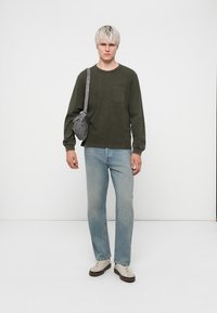 T-shirt à manches longues vert olive avec une poche poitrine, jean droit bleu clair, sac bandoulière gris et baskets beiges avec semelles brunes.