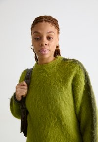 Πράσινο αφράτο πουλόβερ με υφή, στρογγυλή λαιμόκοψη και ελαφρώς σκουρότερες πλευρικές λεπτομέρειες. Το μοντέλο κρατά μια καφέ ζώνη.