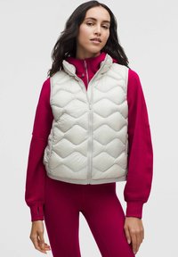 Jasnoszara pikowana kamizelka z wysokim kołnierzem i zamkiem z przodu, noszona na jasnoróżowej bluzce z długim rękawem. Gładka faktura.