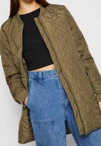 Persoon in een lange olijfgroene gewatteerde jas over een zwart crop-top en hoog-getailleerde blauwe spijkerbroek met zakken.