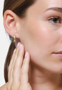 Orecchino a cerchio in oro con una finitura lucida e elegante. Il modello tocca il viso, mettendo in evidenza la curvatura e la superficie riflettente dell'orecchino.