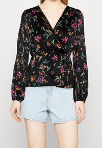 Blouse noire à fleurs avec manches longues, encolure en V, taille cintrée et motifs floraux colorés. Associée à un short en jean bleu clair.