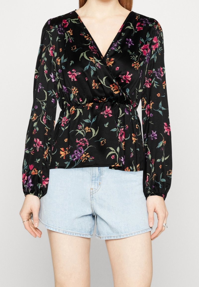 Blouse noire à fleurs avec manches longues, encolure en V, taille cintrée et motifs floraux colorés. Associée à un short en jean bleu clair.