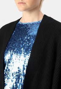 Svart stickad cardigan över en blå paljetttopp, som visar upp glänsande blå paljetter i varierande nyanser och en slät textur.