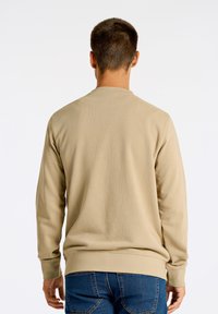 Beige crewneck sweatshirt med långa ärmar, ribbade manschetter och fåll. Materialet verkar mjukt med en slät textur. Bärs med blå jeans.
