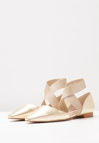 Zapatos planos metálicos dorados con punta afilada, que cuentan con correas elásticas entrecruzadas y un tacón bajo. Textura suave con un detalle mínimo.