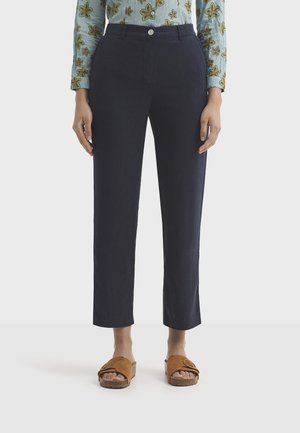 Personne portant un pantalon droit bleu marine, un chemisier à manches longues à fleurs, et des sandales ouvertes en daim marron, debout sur un fond blanc.