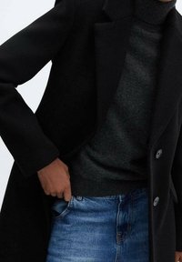 Cappotto di lana nera con colletto a punta, indossato sopra un dolcevita grigio scuro. I jeans in denim blu sono visibili, con un classico design a cinque tasche.