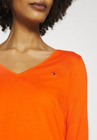 Lys orange V-hals langærmet skjorte lavet af glat stof, med et lille broderet logo på venstre bryst.