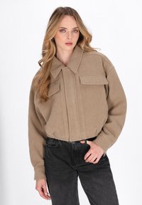 Giacca in misto lana beige con design a colletto, pieghe frontali e due tasche sul petto. Indossata con jeans neri a vita alta.