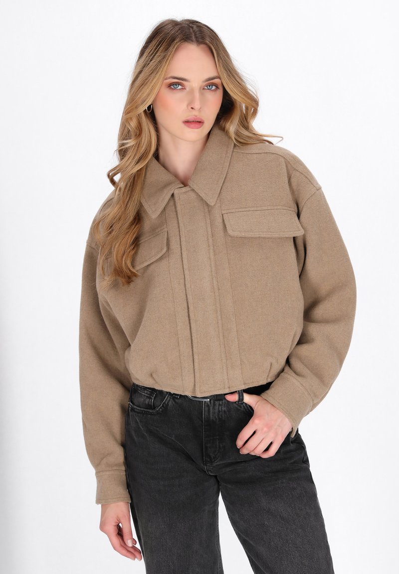 Beige wool-blend jacket with a collared design, front pleats, and two chest pockets. Worn with high-waisted black jeans.