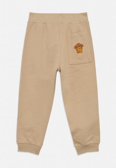 Bézs babagumipántos melegítőnadrág, hátul foltzseb, amelyen sárga és barna hímzett oroszlánfej látható.