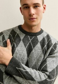 Jonge man in een grijs en zwart ruitpatroon trui, staand tegen een effen lichte muur met één arm over zijn borst gekruist.