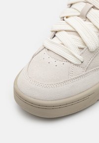 Sneaker bege em camurça com parte superior texturizada, bico redondo e atacadores creme. Sola de borracha grossa e cinza claro com detalhes de costura subtis.