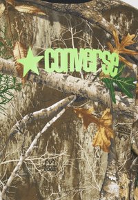 T-shirt met camouflagemotief met een groene "Converse" logo en een ster. Gemaakt van een stof met tinten bruin, groen en gele bladeren, en texturen van boomschors.