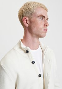 Cardigan lavorato a maglia crema con colletto alto, caratterizzato da quattro bottoni neri, texture a coste e chiusura con zip, indossato sopra una maglietta bianca.