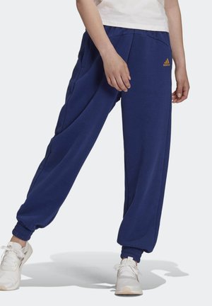 Person iført blå Adidas joggingbukser og hvide sneakers, der står med benene fra hinanden på en ensfarvet baggrund.
