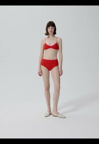 Femme debout, portant un soutien-gorge rouge et une culotte taille haute, ainsi que des chaussures blanches faciles à enfiler, avec une expression neutre sur un fond gris uni.