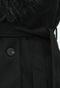 Μαύρο παλτό από μαλλί με κουμπί, ζώνη δεμένη στη μέση και γούνα στο γιακά ορατή στην κορυφή.