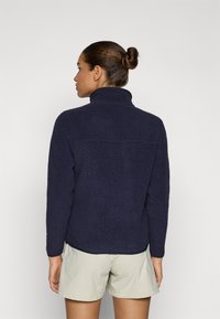 Giacca in pile navy con superficie texture, colletto alto e orlo leggermente curvo, indossata sopra pantaloni corti chiari.