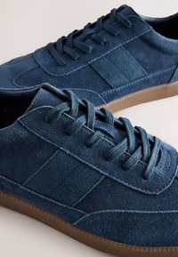 Blå ruskindssneakers med tekstureret overflade, flade snørebånd og gummi sål med diagonalt stribet mønster på siden.