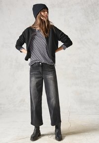 Veste bomber noire texturée, blouse grise à motifs, jeans larges foncés et bottines noires, avec un bonnet en tricot sur la tête.