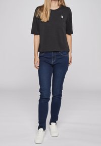 Schwarzes leichtes T-Shirt mit kurzen Ärmeln und kleinem weißen Logo, kombiniert mit dunkelblauen Slim-Fit-Jeans und weißen Sneakers.