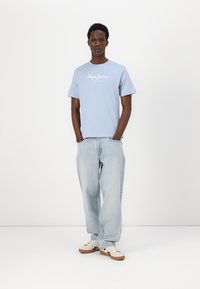 Απαλή μπλε βαμβακερή μπλούζα με κοντά μανίκια και λευκό λογότυπο. Συνδυάζεται με αμάνικες ελαφριές τζιν παντελόνες και μπεζ αθλητικά παπούτσια.