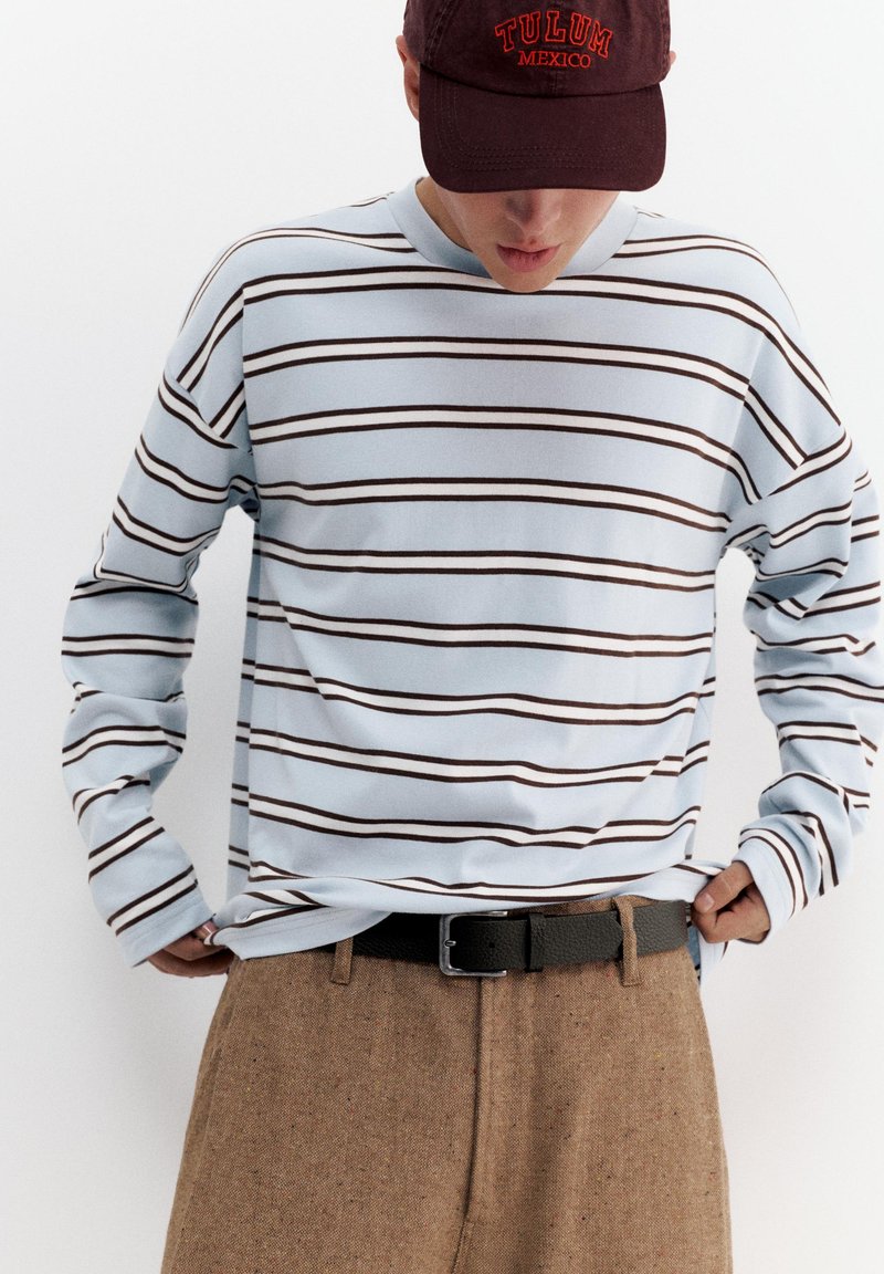 Personne portant une chemise à manches longues bleu clair rayée, un pantalon marron avec une ceinture noire, et une casquette foncée avec l'inscription "TULUM MEXIQUE" en lettres rouges.