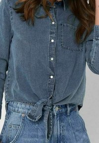 Camicia di denim blu scuro con chiusura a bottoni, colletto, tasca applicata e nodo davanti. Abbinata a pantaloni corti in denim a vita alta.