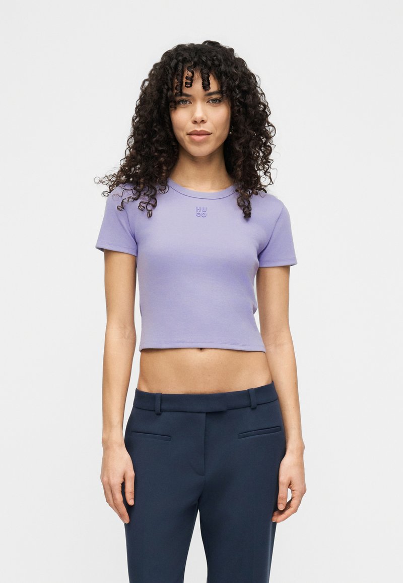 Μωβ croptop μπλουζάκι με κοντά μανίκια, με στρογγυλή λαιμόκοψη και μικρό κεντημένο λογότυπο στο στήθος. Λείο βαμβακερό ύφασμα.