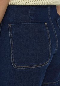 Pantalones de mezclilla azul oscuro con dos bolsillos traseros, acentuados por costuras amarillas en contraste y un acabado suave.