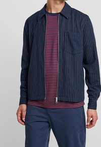 Chaqueta azul marino con cremallera y rayas blancas finas, con cuello puntiagudo y bolsillo en el pecho, combinada con una camisa de rayas burdeos y azul marino.