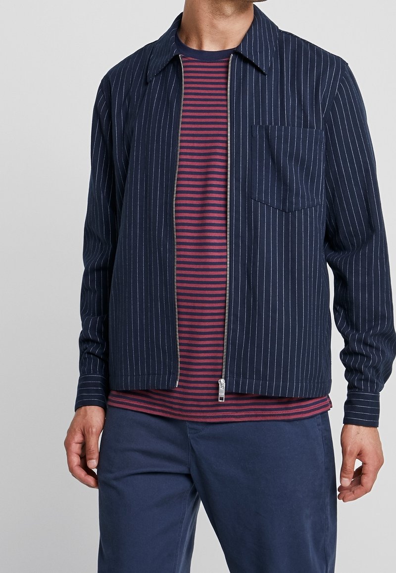 Chaqueta azul marino con cremallera y rayas blancas finas, con cuello puntiagudo y bolsillo en el pecho, combinada con una camisa de rayas burdeos y azul marino.