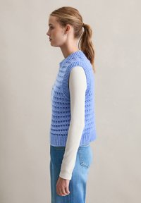 Jasnoniebieski dzianinowy bezrękawnik z teksturowanym wzorem i ściągaczami, noszony na białej koszuli z długim rękawem, w zestawie z niebieskimi dżinsami.