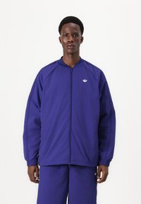 Veste zippée violette avec un col haut, des poignets cintrés et un logo brodé sur la poitrine. Fabriquée en tissu léger et lisse.