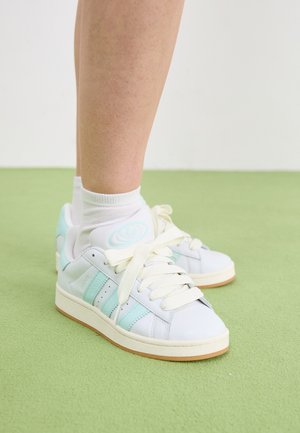 Valged nahast tossud münti aktsentidega, ümmargune logo ja kreemivärvi paelad; tekstureeritud kummitald; koos valgete sokkidega rohelisel põrandal.