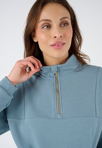 Felpa zip blu chiaro con colletto alto, caratterizzata da una cerniera dorata e dettagli delle cuciture sulla parte anteriore. Tessuto morbido e dalle texture.