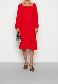 Červené šaty s dlouhým rukávem a hranatým výstřihem, s detailem na zavázání, obsahující vertikální plisování. Spárováno s černými sandály na řemínky a pruhovanou kabelkou.